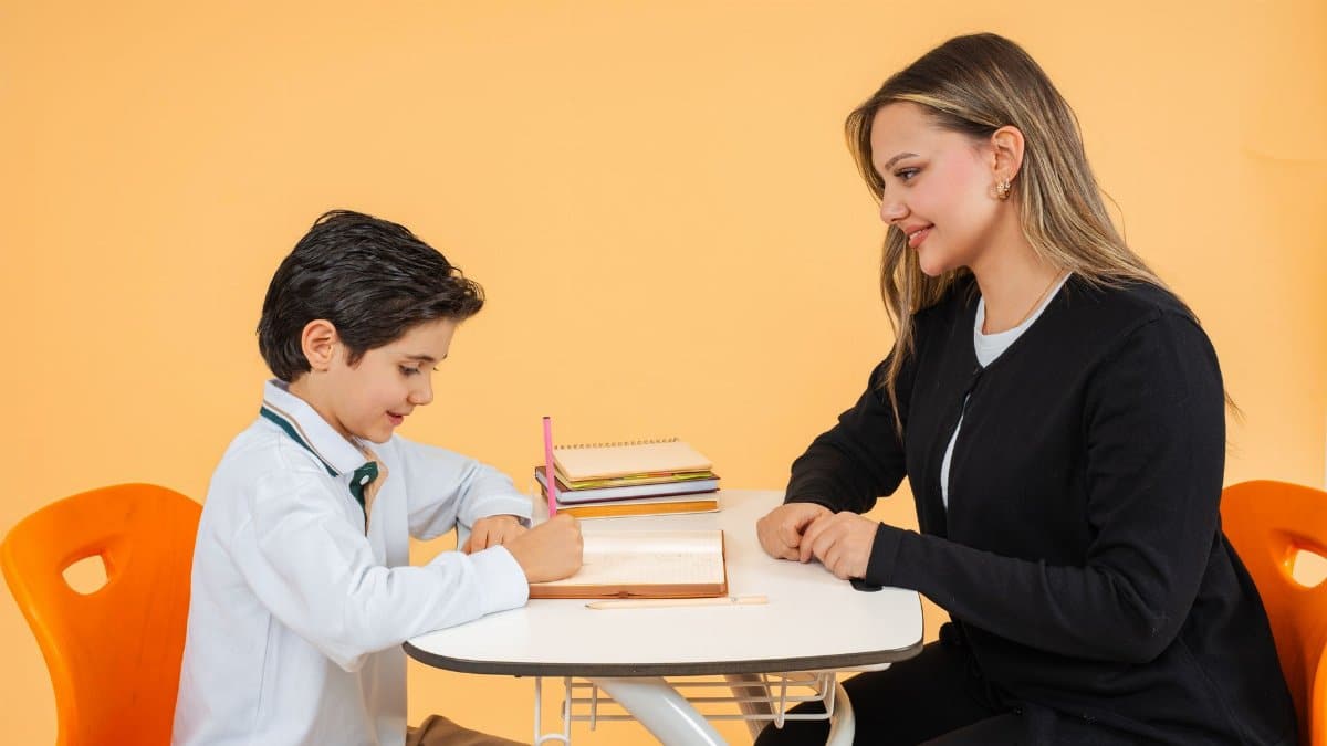 A teacher helps a student with schoolwork at a desk in a bright classroom.