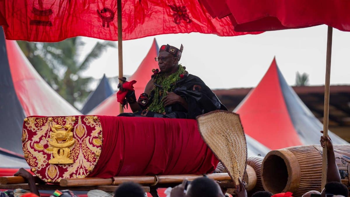 A king in ceremonial attire sits under a vibrant red canopy during a traditional African festivity.