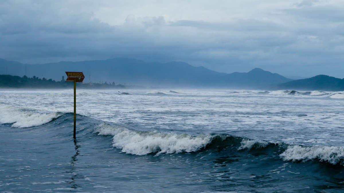 Cloudy coastal scene with warning sign and waves crashing against the shore, conveying sea caution.