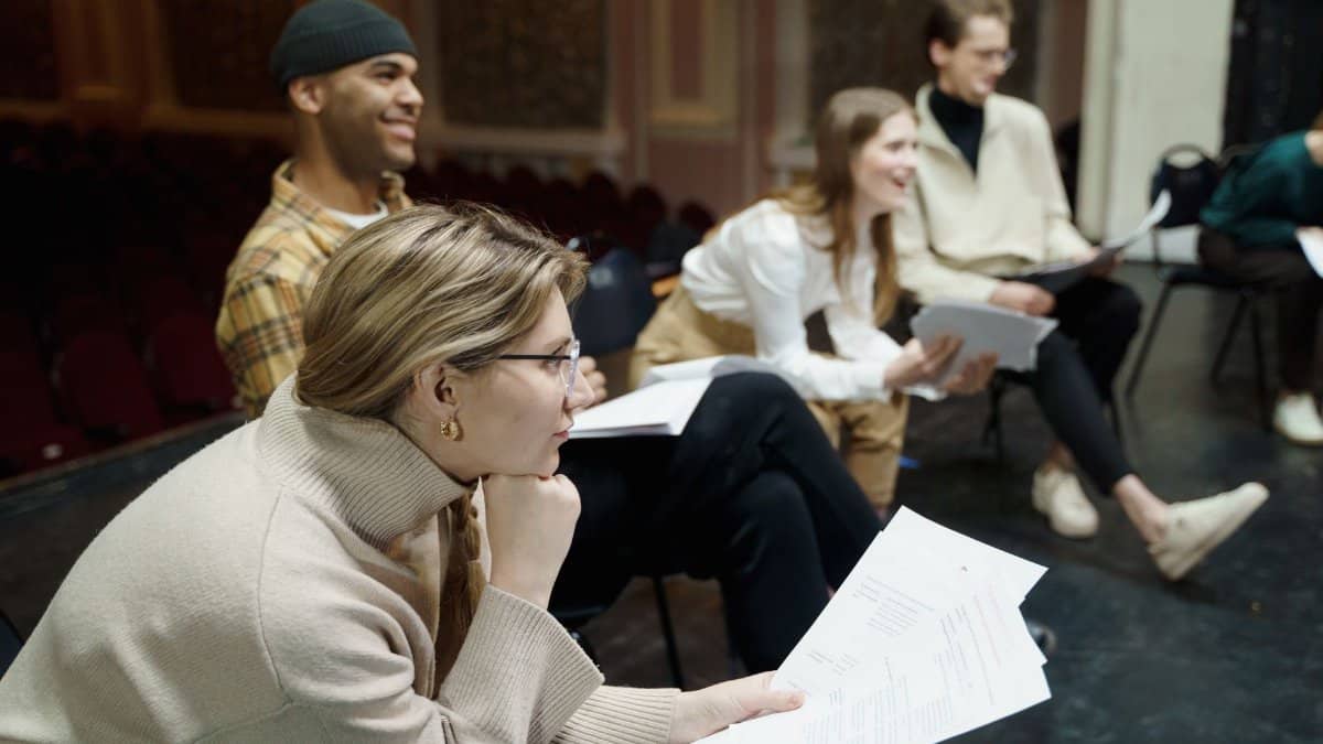 Actors rehearsing a play on stage, holding scripts and discussing roles in a theater setting.
