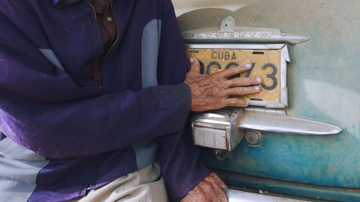 A senior adult touching a vintage Cuban license plate on a classic car, reflecting heritage.