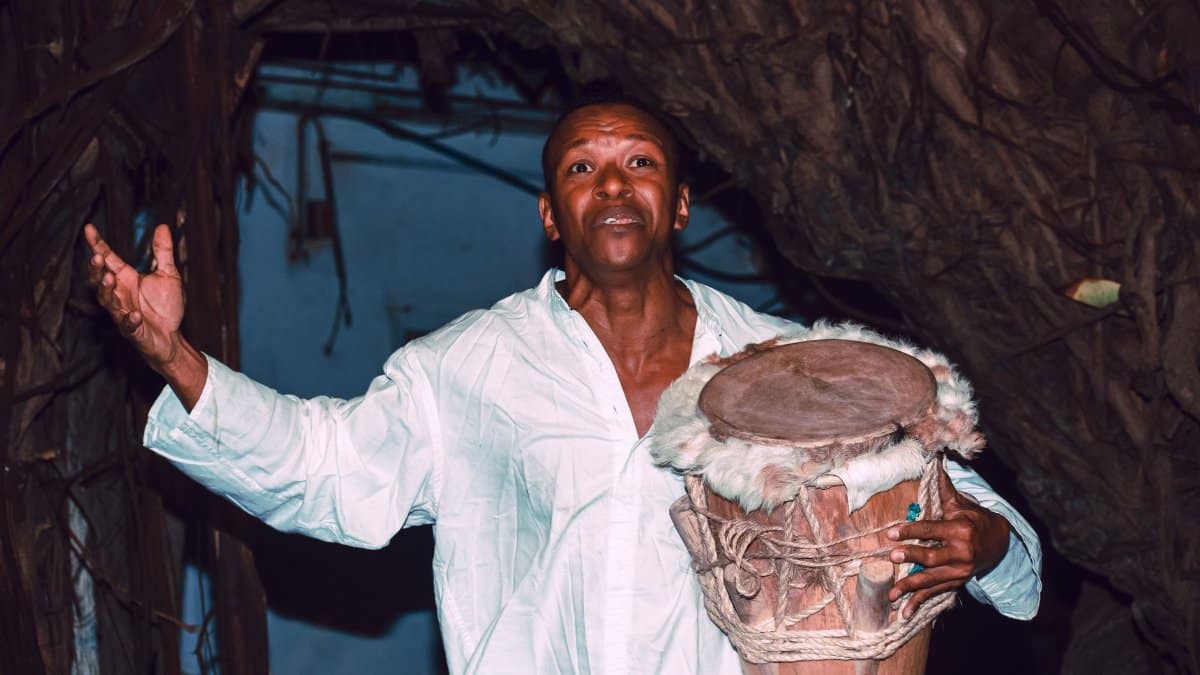 A man in a white shirt holds a traditional drum outdoors, expressing emotion.