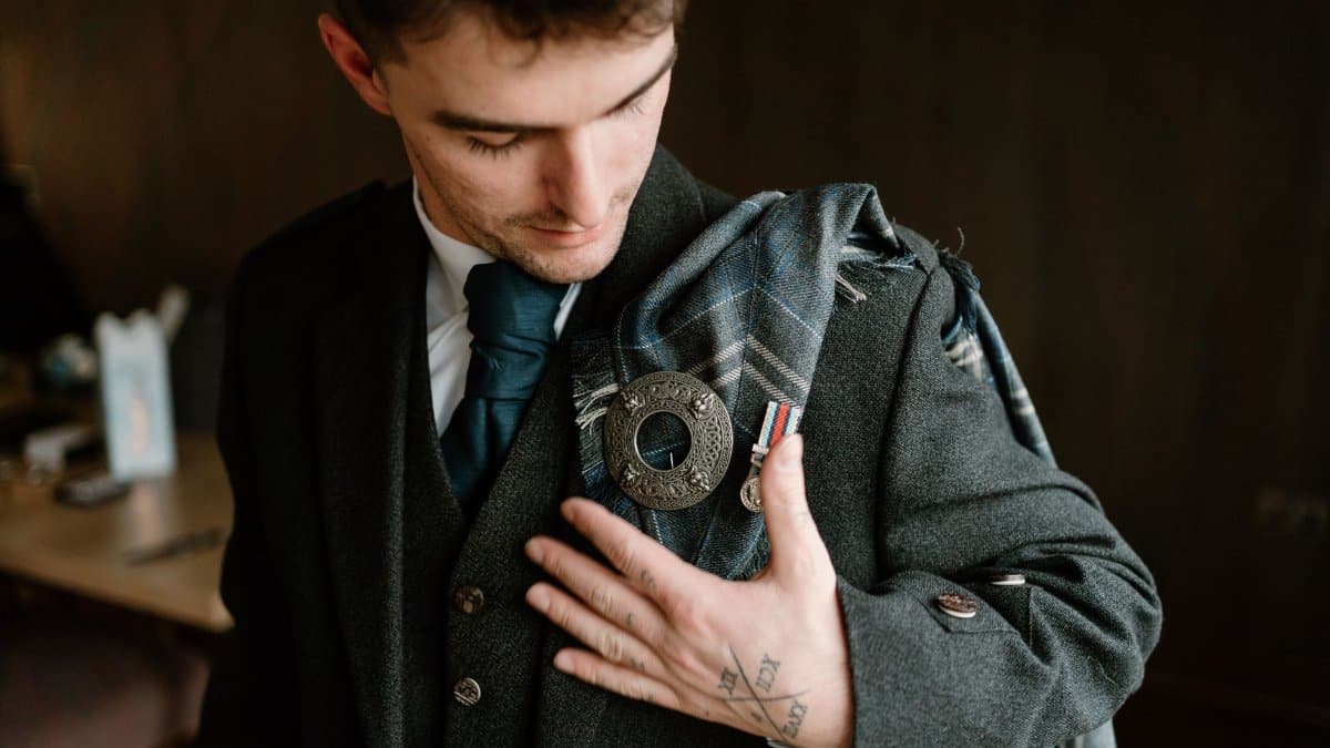Groom in traditional Scottish attire adjusting his tartan brooch indoors before wedding ceremony.