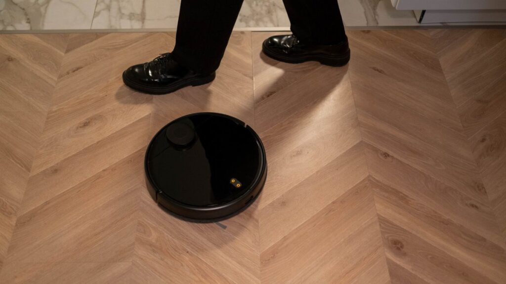 A modern robot vacuum cleaner navigating wooden floors while a person in black shoes walks past.
