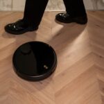 A modern robot vacuum cleaner navigating wooden floors while a person in black shoes walks past.