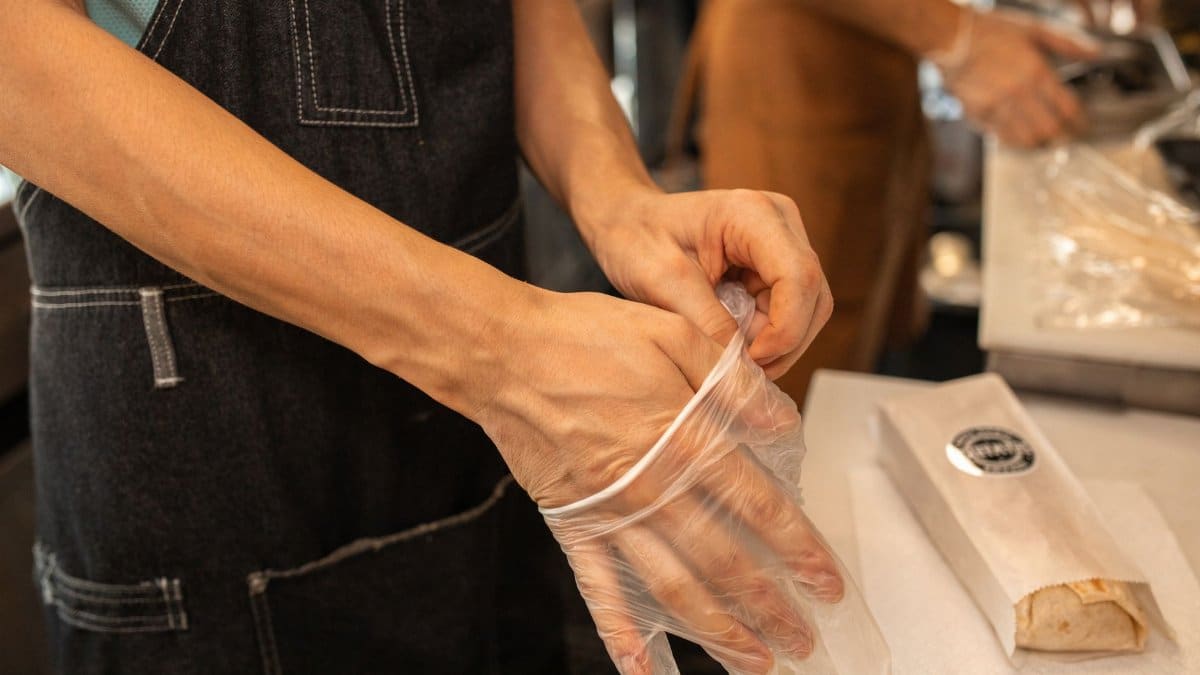 A person wearing an apron puts on plastic gloves in a kitchen environment, preparing for food handling.