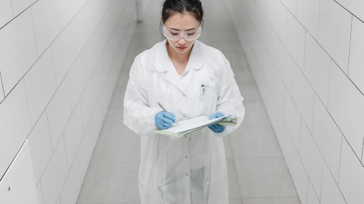A female scientist wearing safety goggles and a lab coat taking notes in a laboratory corridor.