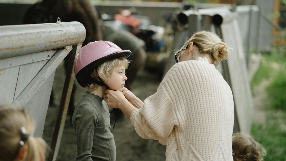 Mother helping child with a pink helmet in a farm setting, emphasizing care and safety.