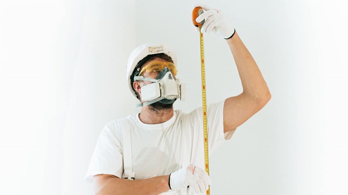A construction worker wearing safety equipment measures with a tape against a light background.