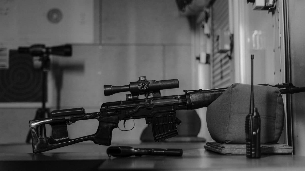Monochrome image of a sniper rifle on a table indoors with a walkie talkie and scope.