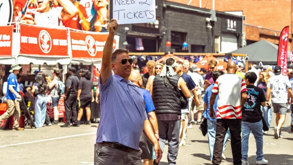 Casual man holding 'Need 2 Tickets' sign amidst crowd at Seattle event.