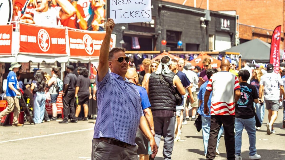 Casual man holding 'Need 2 Tickets' sign amidst crowd at Seattle event.