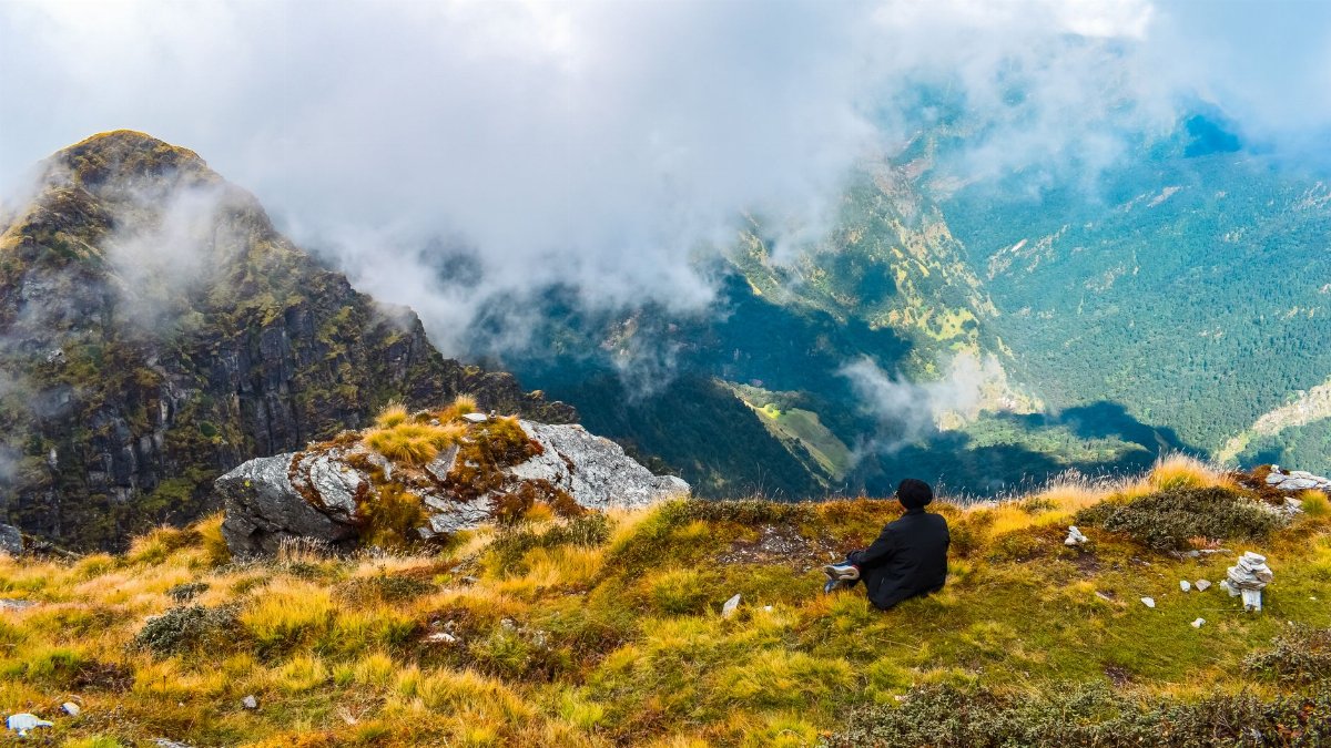 Person sitting on a grassy mountain cliff overlooking foggy valley. Adventure and solitude in nature.