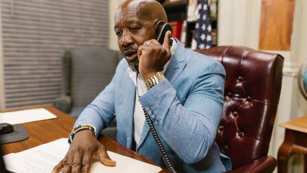 African American man in a blue suit speaking on the phone in a professional office setting.