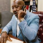 African American man in a blue suit speaking on the phone in a professional office setting.
