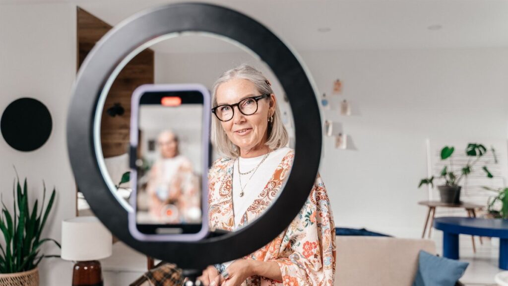 Elegant senior woman recording a video using a smartphone and ring light in a stylish home setting.