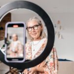 Elegant senior woman recording a video using a smartphone and ring light in a stylish home setting.