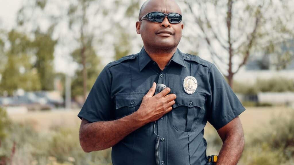 Confident police officer in uniform outdoors, hand on badge, sunny park setting.