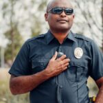 Confident police officer in uniform outdoors, hand on badge, sunny park setting.
