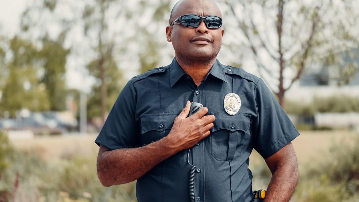 Confident police officer in uniform outdoors, hand on badge, sunny park setting.