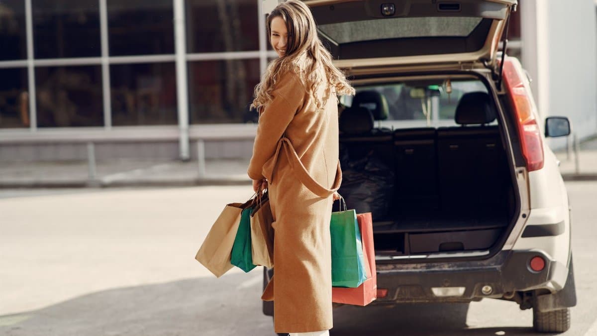 Full length of cheerful female looking over shoulder while standing near opened trunk of car on parking of shopping mall