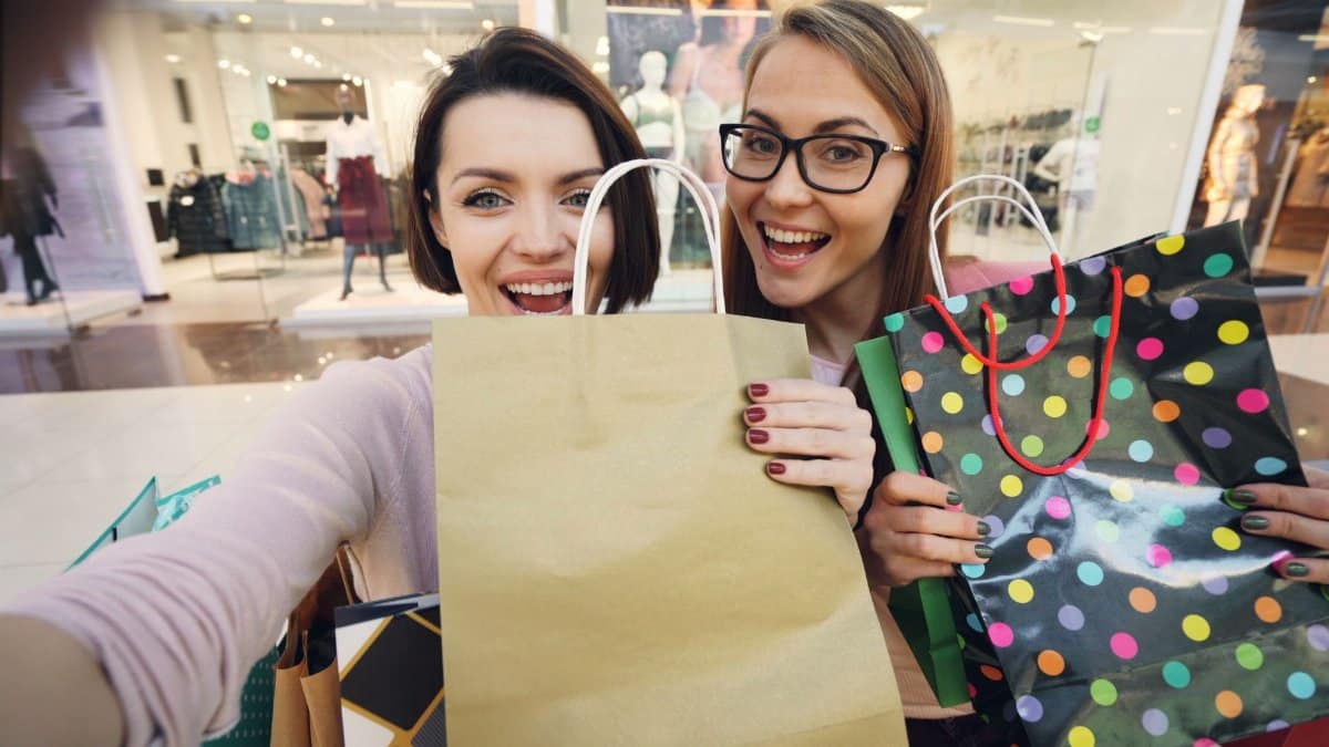 Two women smiling with shopping bags in a mall environment.