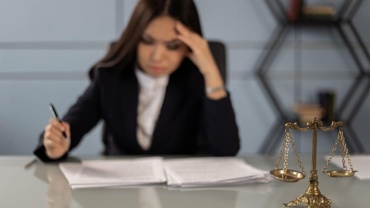 A fatigued female lawyer reviewing documents with a justice scale nearby, emphasizing stress in legal work.