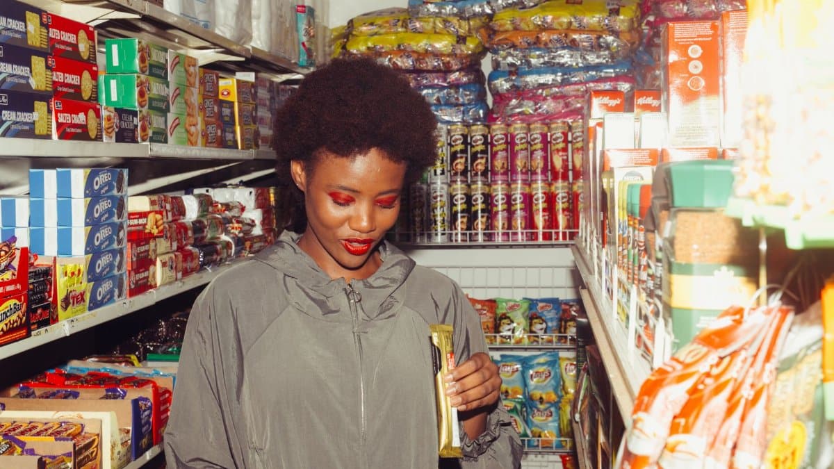 Adult black female buyer in casual outerwear with bright makeup holding chocolate product while standing in supermarket and choosing goods for purchase