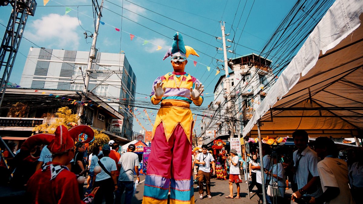 Colorful street performer on stilts entertains a festive crowd outdoors under a bright sky.