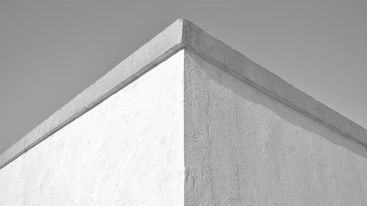 A minimalist photograph of a concrete wall corner under a clear sky, emphasizing texture and form.