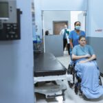 A nurse helps a patient in a wheelchair down a hospital corridor, reflecting care and medical professionalism.