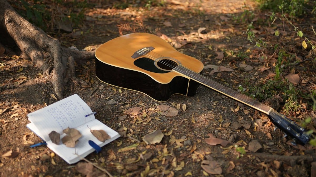 An acoustic guitar rests on forest ground beside an open notebook, capturing the essence of music creation in nature.