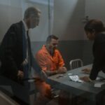 A tense interrogation in progress with a handcuffed inmate and two officers in a prison room.