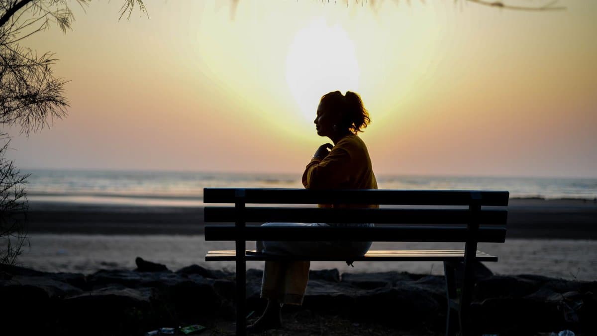 A silhouette of a woman sitting on a bench by the beach during sunrise, capturing solitude and contemplation.