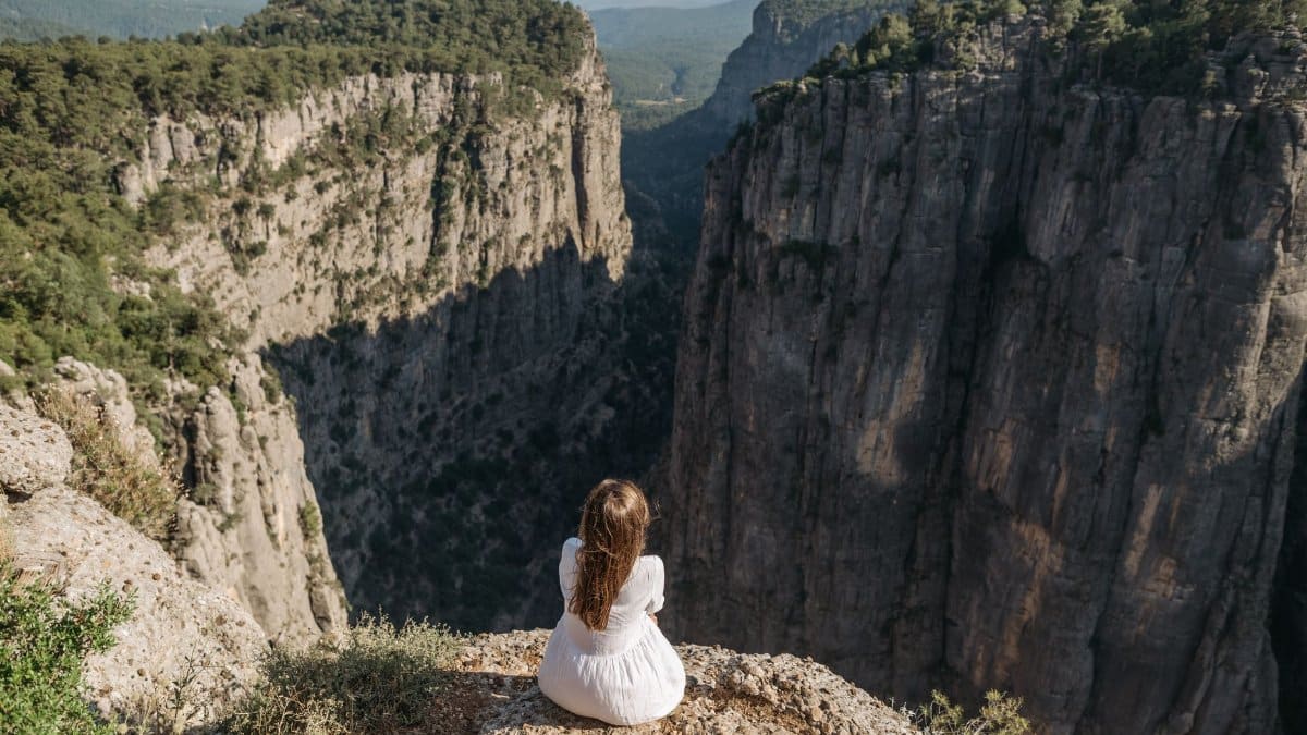 A woman in a white dress sitting alone, overlooking a deep canyon in a mountainous area.