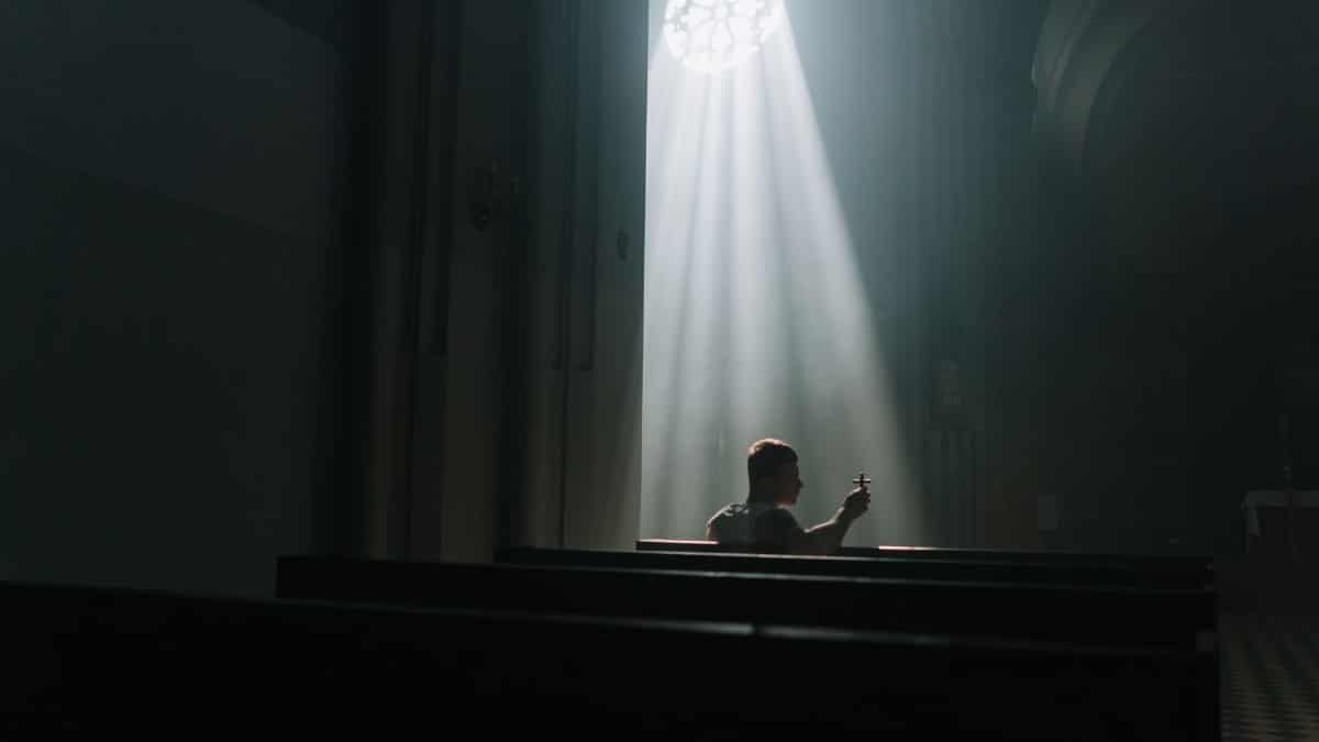 A person sits praying inside a church, illuminated by rays of sunlight through a window.