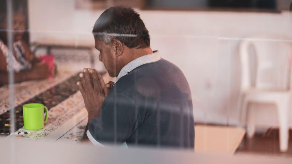 A man praying indoors at a table with a warm, comforting atmosphere.