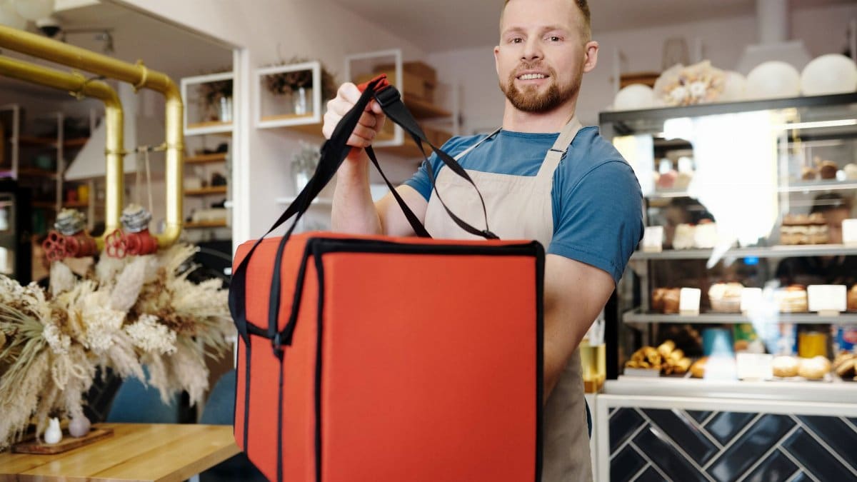 Smiling man in an apron holding a delivery bag in a cozy cafe setting, ready for service.