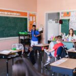 Children in a classroom with a teacher on the first day of school, chalkboard with "Bring Your Thinking Cap" message.