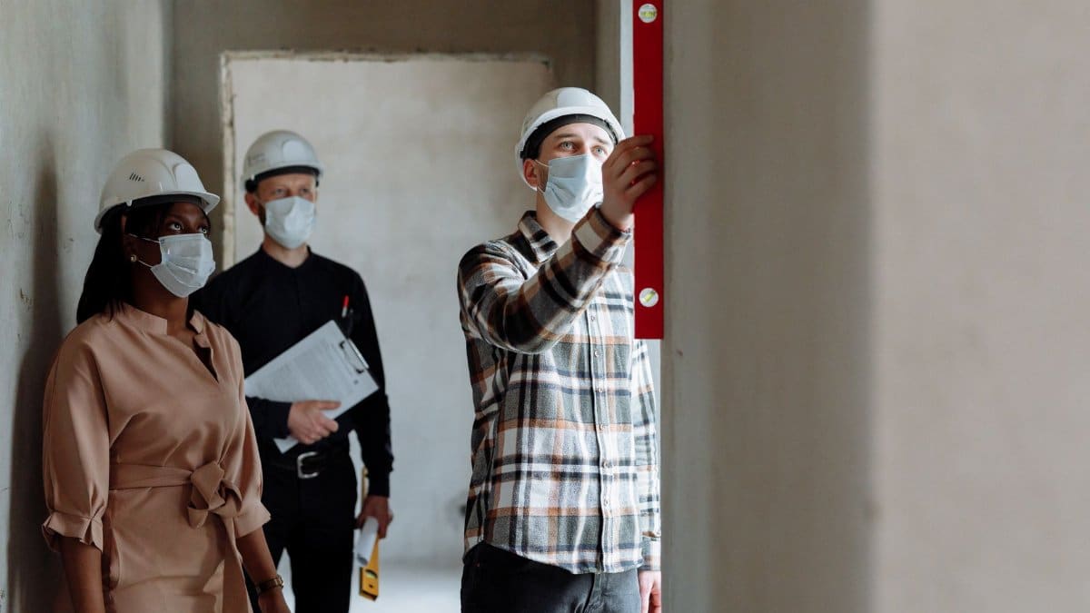 Team of construction workers in hard hats and masks inspecting indoors using a level.