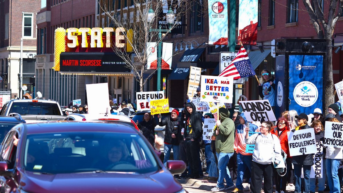 Protestors gather outside the State Theatre holding signs with various messages in a bustling city scene.