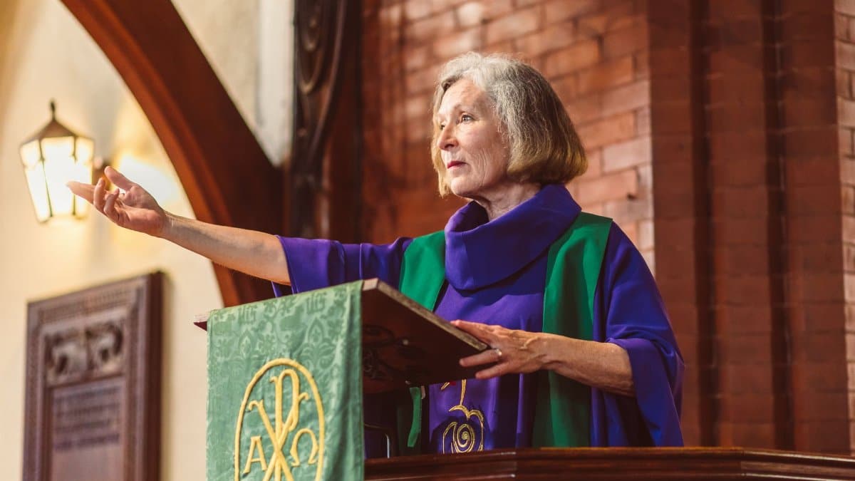 Elderly female pastor giving a sermon from a lectern in a church setting, arm extended.