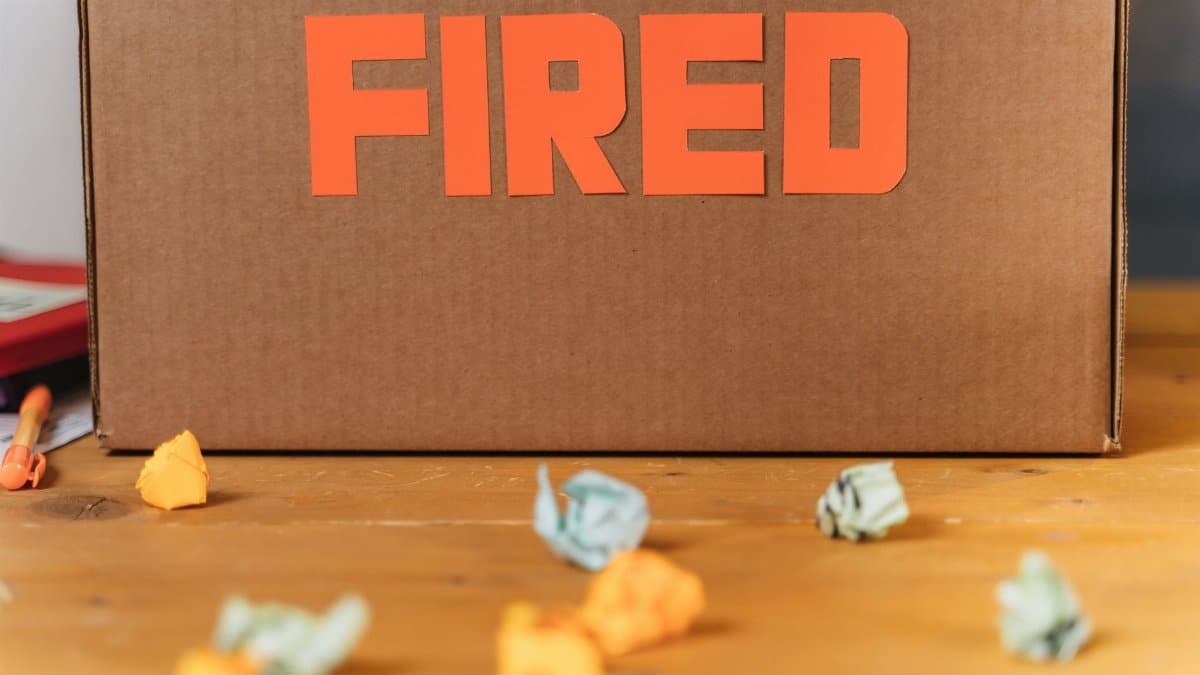 Close-up of a cardboard box labeled FIRED on a wooden table with crumpled papers.