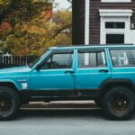 Bright blue vintage Jeep Cherokee parked by a rustic building on a tree-lined street.
