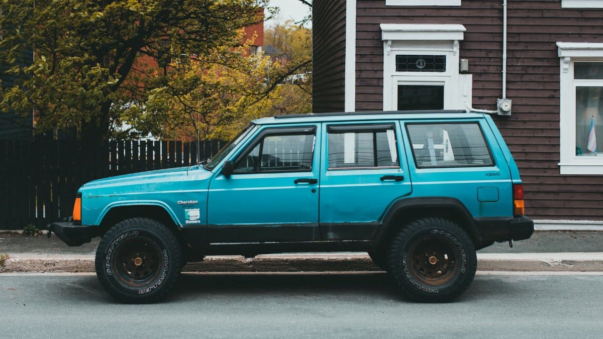 Bright blue vintage Jeep Cherokee parked by a rustic building on a tree-lined street.