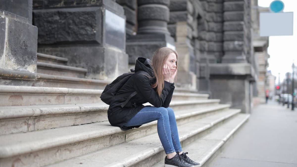 A young woman in casual wear sits alone on stone steps outside a historic building.