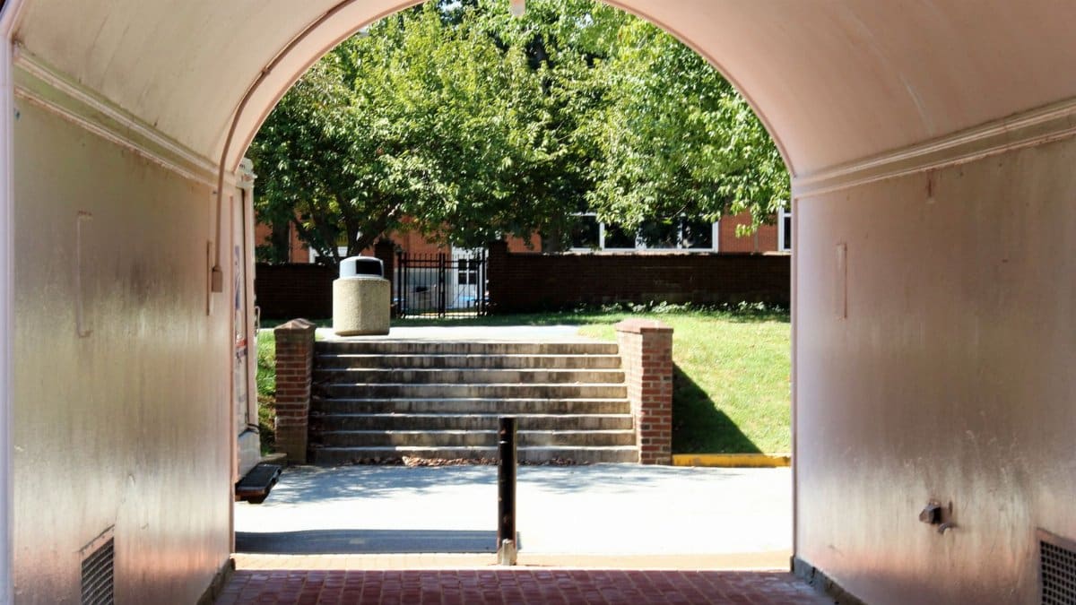 View through an archway to a sunlit area with steps, trees, and buildings in Newark, DE.