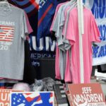 A stall displaying Trump 2020 merchandise including shirts and signs at an outdoor market.