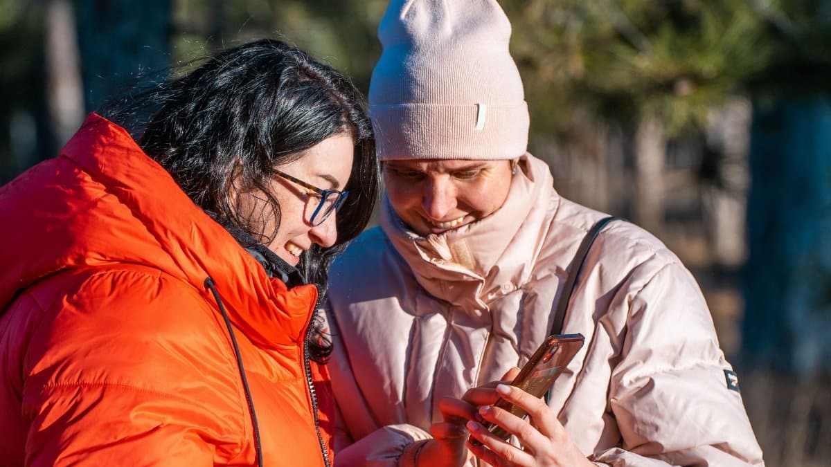Two friends in winter jackets enjoy looking at their smartphones outdoors in bright daylight.