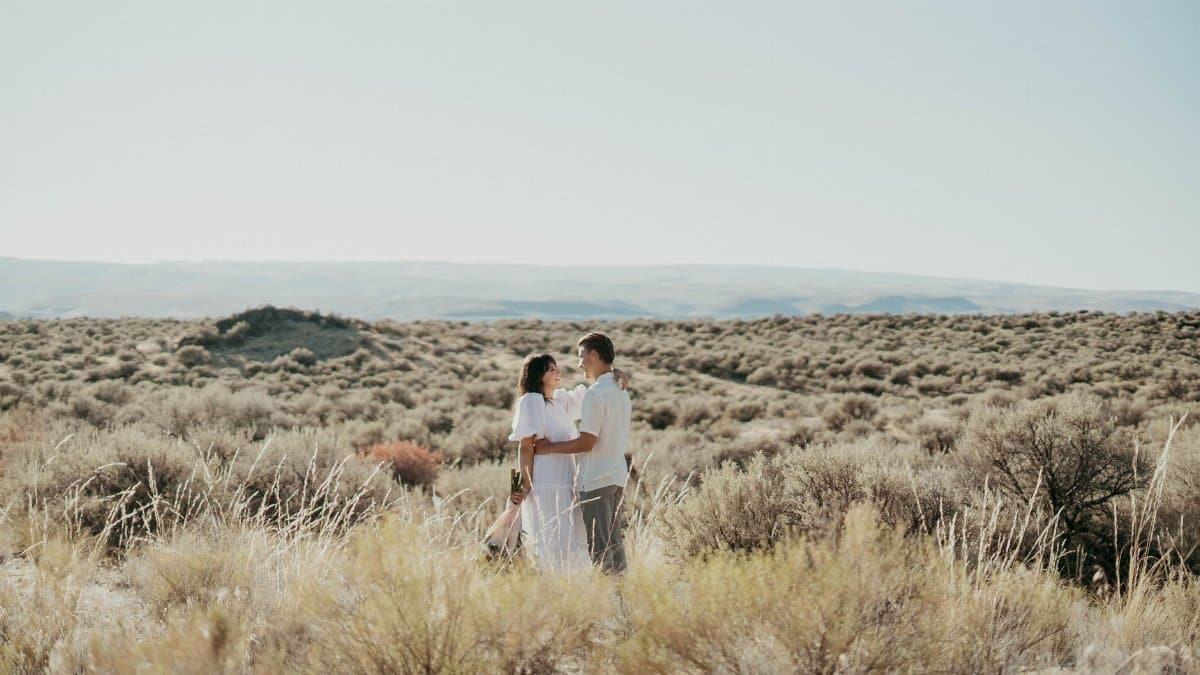 Cheerful couple in stylish clothes hugging while standing in field with dry bushes in countryside and looking at each other